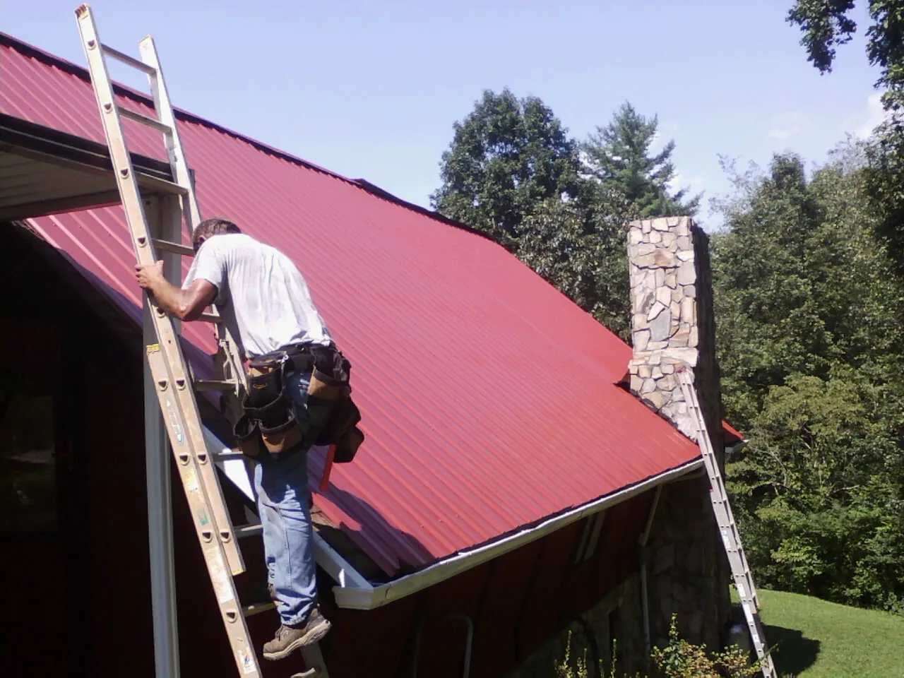 Roofer on a ladder beside a red metal roof and stone chimney — Soffit & Fascia Repair by Roofing Brothers in Mayslick